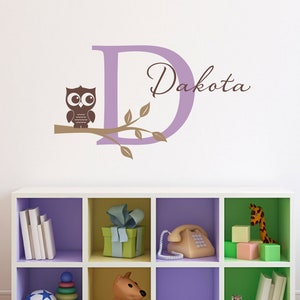 Puede incluir: Una letra D morada con el nombre Dakota escrito en cursiva marrón. Un búho marrón está posado en una rama con hojas verdes. La letra y el búho están en una pared blanca. Debajo de la calcomanía de pared hay una estantería blanca con seis compartimentos. La estantería está pintada en diferentes colores y tiene varios juguetes y libros dentro.