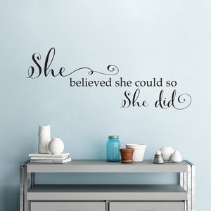May include: A light blue wall with a black vinyl decal that reads "She believed she could so she did."  A gray dresser with two drawers sits in front of the wall.  The dresser has a variety of decorative items on top, including a white vase, a blue glass jar, and a white ceramic pot.