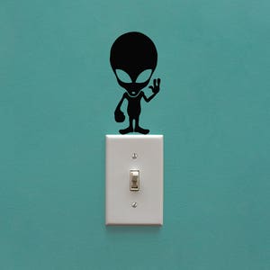May include: A black alien sticker with a friendly wave, placed on a white light switch against a teal wall.