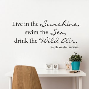 May include: A white wall with a black vinyl decal quote that reads "Live in the Sunshine, swim the Sea, drink the Wild Air." The quote is attributed to Ralph Waldo Emerson. The wall is behind a white desk with a wooden chair, two white heart-shaped decorative objects, a glass jar of pencils, and a blue planter with a green plant.