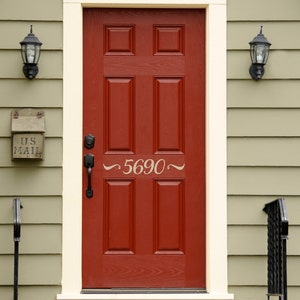 May include: A red front door with a white frame and a white house number plaque with the number 5690. The door has six panels and a black door handle. There is a black mailbox with the words "U.S. MAIL" on it to the left of the door.