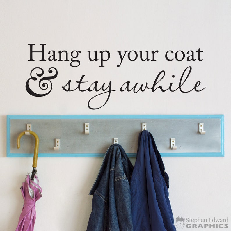 May include: A white wall with a black vinyl decal that reads "Hang up your coat & stay awhile". A wooden coat rack with a light blue trim is mounted below the decal. Two jackets and an umbrella hang on the rack.