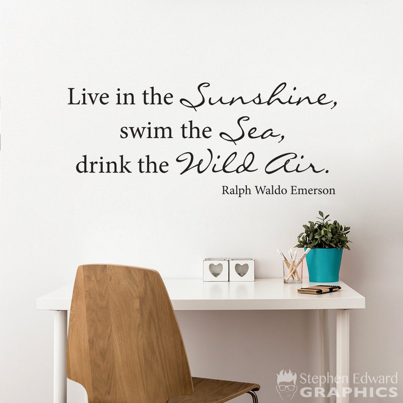 May include: A white wall with a black vinyl decal quote that reads "Live in the Sunshine, swim the Sea, drink the Wild Air." The quote is attributed to Ralph Waldo Emerson. The wall is behind a white desk with a wooden chair, two white heart-shaped decorative objects, a glass jar of pencils, and a blue planter with a green plant.