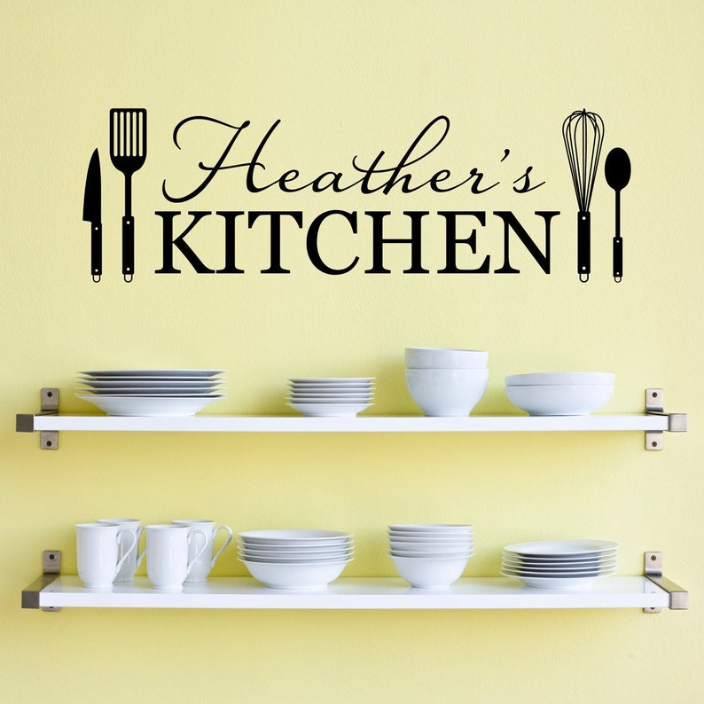 May include: White kitchen shelves with white dishes and bowls. The wall behind the shelves has a black vinyl decal that says "Heather's Kitchen" with kitchen utensil silhouettes.