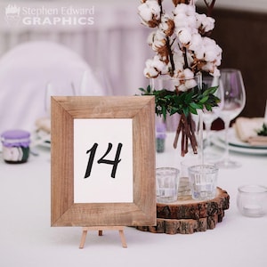 May include: A wooden frame with a white card inside, displaying the number "14" in black calligraphy. The frame is on a small wooden easel and sits on a white tablecloth. A vase with cotton branches and greenery is in the background.