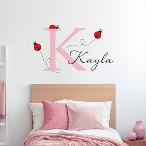 Könnte beinhalten: Rosaer Buchstabe K mit dem Namen Kayla darunter in Kursivschrift. Drei rote Marienkäfer mit schwarzen Punkten fliegen um den Buchstaben, jeder mit einer gepunkteten Linie, die hinter ihm herzieht.