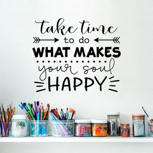 Puede incluir: Una pared blanca con letras negras que dicen "Tómate el tiempo para hacer lo que hace feliz a tu alma". Hay flechas a cada lado del texto. Debajo del texto hay frascos llenos de pinceles coloridos.