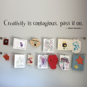 May include: A white wall with the quote "Creativity is contagious, pass it on. - Albert Einstein -" in black lettering.  The wall has a wire strung across it with various children's artwork hanging from it. The artwork includes drawings of fish, hearts, a boat, and an octopus.