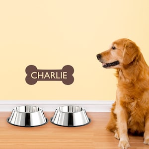 May include: Two stainless steel dog bowls on a wooden floor. A golden retriever dog is sitting to the right of the bowls, looking at the bowls. A brown bone-shaped sign with the name "CHARLIE" is on the wall behind the bowls.