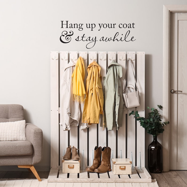 May include: White wooden coat rack with hooks and a sign that reads "Hang up your coat & stay awhile". There are four coats hanging on the rack, including a yellow jacket, a white jacket, a green jacket, and a scarf. There are also two pairs of brown boots and a brown purse on the rack.