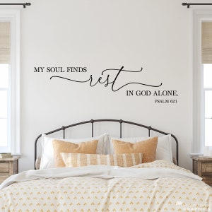 May include: A white wall with a black vinyl decal that reads "My soul finds rest in God alone. Psalm 62:1". The decal is above a bed with a black metal headboard and a white comforter with a yellow and white patterned duvet cover.