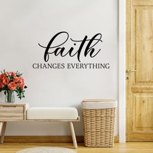 Puede incluir: Una pared blanca con una calcomanía de vinilo negro que dice "la fe lo cambia todo". Un cesto de ropa de mimbre y un banco de madera están en primer plano.