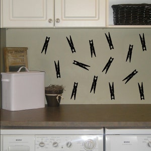May include: Black clothespin wall decals, arranged in a random pattern on a light brown wall. The decals are in a laundry room setting with a white washing machine and dryer in the foreground.