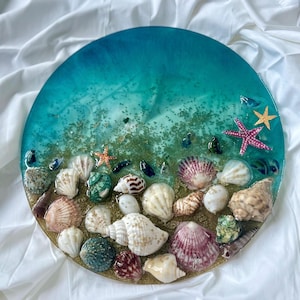 Op de afbeelding: Een rond kunstwerk van hars met een kleurverloop van diep blauwgroen naar licht turkoois, ingelegd met diverse schelpen, waaronder witte, bruine en paarse varianten, samen met twee zeesterren en verspreide gouden glitterdeeltjes.