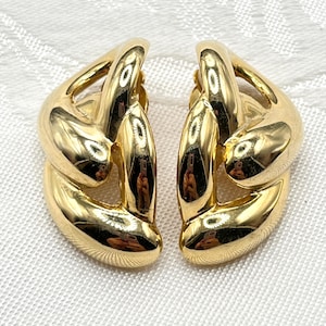 May include: A pair of gold-toned, chunky clip-on earrings. The earrings have a linked chain design, with a polished, reflective surface. The earrings are set against a white, textured background.