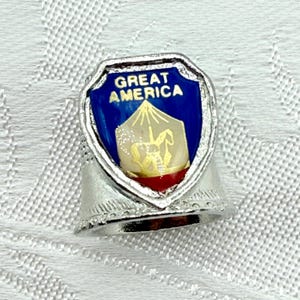 May include: A silver thimble with a shield-shaped emblem. The emblem features the words "GREAT AMERICA" above a gold and white image on a blue background, with a red accent at the bottom. The thimble has a decorative band.