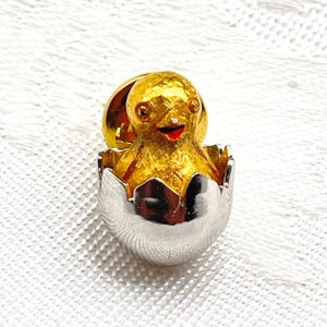 May include: A gold-toned textured metal chick pin emerging from a polished silver-toned metal eggshell. The chick features small dark eyes and a bright red beak. This small decorative accessory is designed with a pin backing for secure attachment.