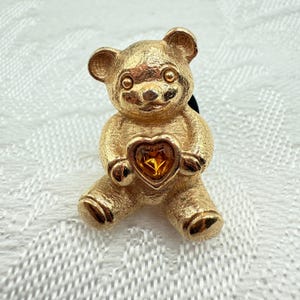 May include: Gold-toned teddy bear brooch holding a heart-shaped gem. The bear has detailed fur texture and is sitting with its legs extended. The heart gem is a warm amber color.