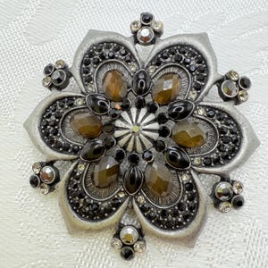 May include: A silver-toned flower-shaped brooch with a detailed design. The brooch features brown and black gemstones, with smaller clear and black stones around the edges. The center has a starburst design.