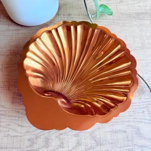 May include: A copper-colored, shell-shaped decorative dish. The dish has a scalloped edge and a textured, ridged interior. The metallic finish reflects light, creating highlights and shadows. The dish is likely used for serving or display.
