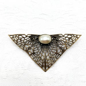 May include: A vintage-style brooch with a triangular, openwork design in a bronze tone. The brooch features a central, oval-shaped faux pearl. The intricate metalwork creates a delicate, lacy pattern, suitable for adding a touch of elegance to any outfit.