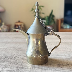 May include: A vintage brass coffee pot with a curved spout and handle. The pot features an etched palm tree design on the body and a hinged lid. The pot is made of brass and has a tarnished finish.