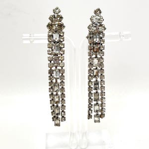 May include: A pair of silver-toned, dangling earrings. Each earring features a cluster of clear, faceted crystals at the top, with multiple strands of rectangular and square crystals cascading downwards. The earrings are displayed on a clear stand against a white background.