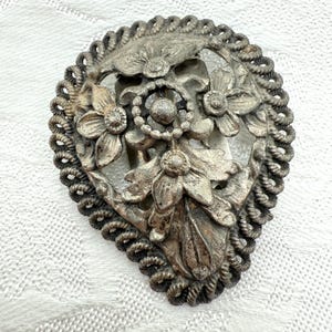 May include: Antique silver-toned metal brooch with floral and leaf design. The brooch features three-dimensional flowers and leaves within a decorative rope-like border. The clasp is visible on the back. A vintage accessory.