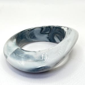 May include: Oval-shaped bangle bracelet in shades of white, gray, and black. The marbled design creates a swirling pattern throughout the bracelet. The bracelet has a smooth, glossy finish.