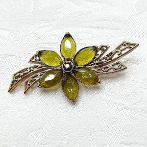 May include: A gold-toned metal brooch featuring five marquise-cut olive green glass stones arranged in a flower shape. The center of the flower is a small, dark red circular stone. The brooch includes decorative filigree metal leaves on the sides.