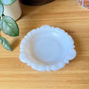May include: A small, white, decorative dish with a scalloped edge. The dish is made of opaque glass and sits on a wooden surface. The dish has a shallow, round interior.