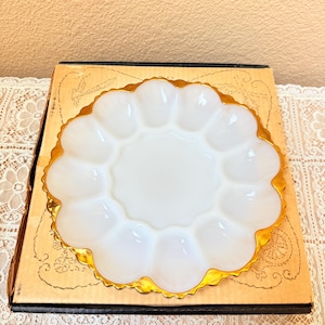 May include: A white, scalloped egg serving dish with a gold rim. The dish has 12 individual egg compartments and a central well. The dish is sitting on a vintage box with a decorative design.