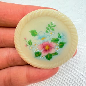 May include: A round, off-white decorative button with a floral design. The center features a pink and yellow flower surrounded by white and blue flowers and green leaves. The button has a rope-like border.