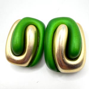 May include: A pair of earrings featuring a smooth, matte green U-shaped component interlocked with a polished gold-tone metal U-shaped component. The design creates a continuous, rounded rectangular loop shape with contrasting textures and colors.