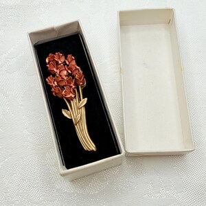 May include: A vintage brooch in a gift box. The brooch features a bouquet of red flowers with gold-colored stems and leaves. The brooch is set against a black velvet background inside a white box.