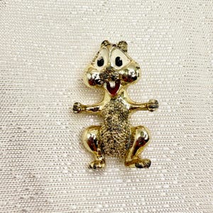 May include: A gold-colored squirrel brooch with black eyes and a red mouth. The squirrel is standing on its hind legs with its arms outstretched.