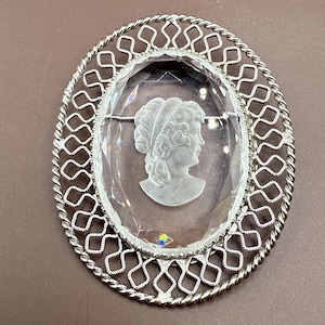 May include: Oval-shaped cameo brooch with a clear, faceted center featuring a profile of a woman. The cameo is set in a silver-toned metal frame with a decorative openwork border. The brooch has a pin closure on the back.