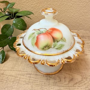 May include: A white, vintage-style covered dish with a gold trim and a pedestal base. The lid features a hand-painted design of two apples with green leaves. The dish is likely made of porcelain or milk glass.