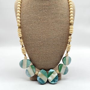 May include: A statement necklace featuring a mix of materials and textures. The necklace has a strand of cream-colored wooden beads, with alternating disc-shaped beads and gold-colored spacers. The focal point includes large, oval-shaped pendants in shades of teal, white, and green.