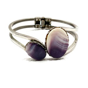 May include: A silver-toned bracelet with two oval, polished stones. The stones are a gradient of purple and white, set in a silver frame. The bracelet has a hinged clasp and a curved design, suitable for jewelry.