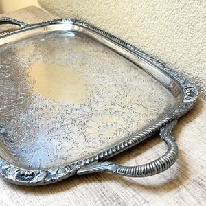 May include: Silver-plated serving tray with ornate floral and scrollwork detailing. The rectangular tray features curved handles and a decorative border. The tray is displayed on a light-colored wooden surface.