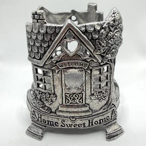 May include: A silver-toned metal candle holder shaped like a small house with a textured roof, windows, and a door. The front features the word Welcome above the door and the phrase Home Sweet Home embossed on the base with three sturdy feet.