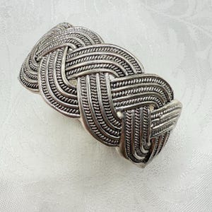 May include: A silver-toned cuff bracelet with an intricate woven design. The bracelet features a series of interwoven bands, creating a textured, knotted appearance. The metal has a slightly aged look, enhancing the detail of the woven pattern. The bracelet is a statement piece.