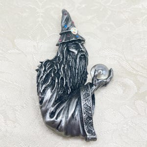 May include: A silver-colored wizard figurine with a long beard and a pointed hat adorned with colorful gems. The wizard holds a clear crystal ball. The figurine has intricate details and a medieval fantasy theme.