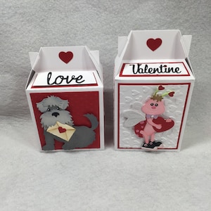May include: Two small white boxes with red accents. The box on the left has a gray dog with a letter and the word "love" on the front. The box on the right has a pink fairy with wings and the word "Valentine" on the front.