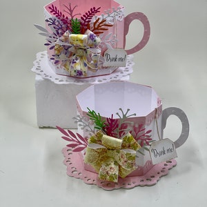 May include: Two pink paper teacups with white lace doilies, decorated with floral paper bows, green and pink paper leaves, and a white tag that says "Drink me!"