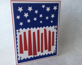 May include: A handmade card with a blue and white American flag design. The flag is made of paper and has white stars on a blue background. The card also features a red, white, and blue design that resembles a cityscape.