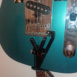 May include: A teal electric guitar with a leopard print strap hanging on a wall mount. The guitar has a chrome bridge and a black pickup.