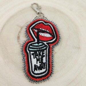 May include: A beaded keychain with a red lip design and a coffee cup with the text "TAKE ME AWAY". The keychain has a silver clasp and is outlined with red and grey beads. The design is reminiscent of a coffee shop.