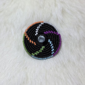 May include: A round, beaded brooch with a black base and colorful accents. The brooch features a central, faceted, silver-colored gem. The design includes orange, green, purple, and blue beaded lines radiating from the center.